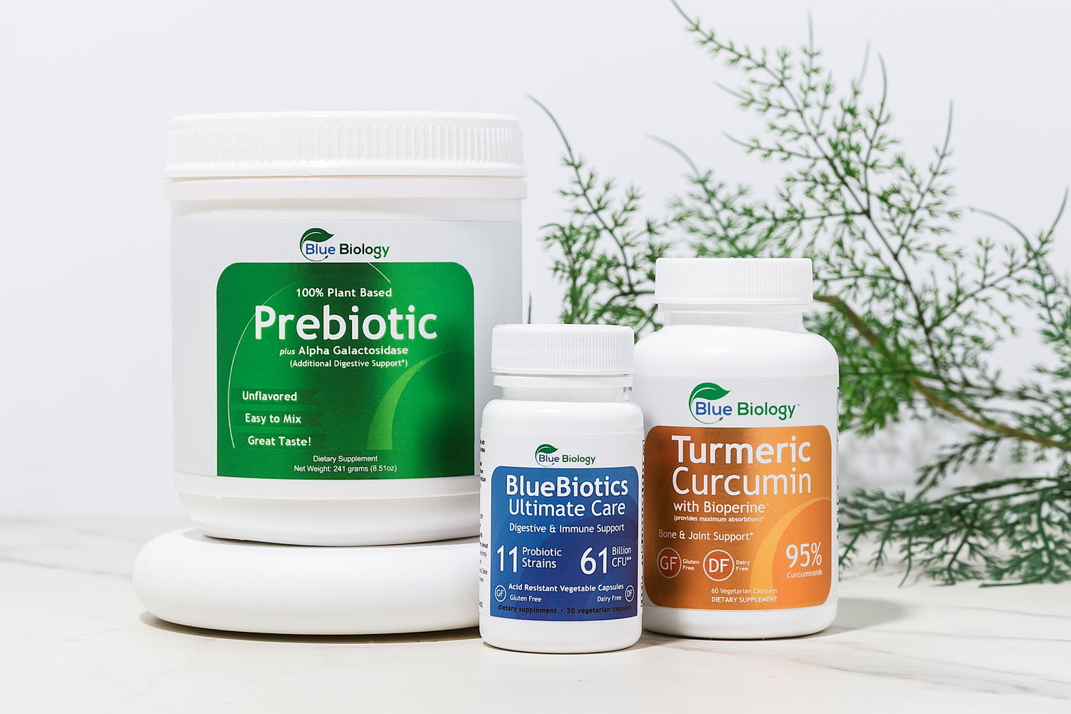 Supplement products including prebiotic powder, turmeric curcumin, and other health supplements on a white surface with a plant in the background.